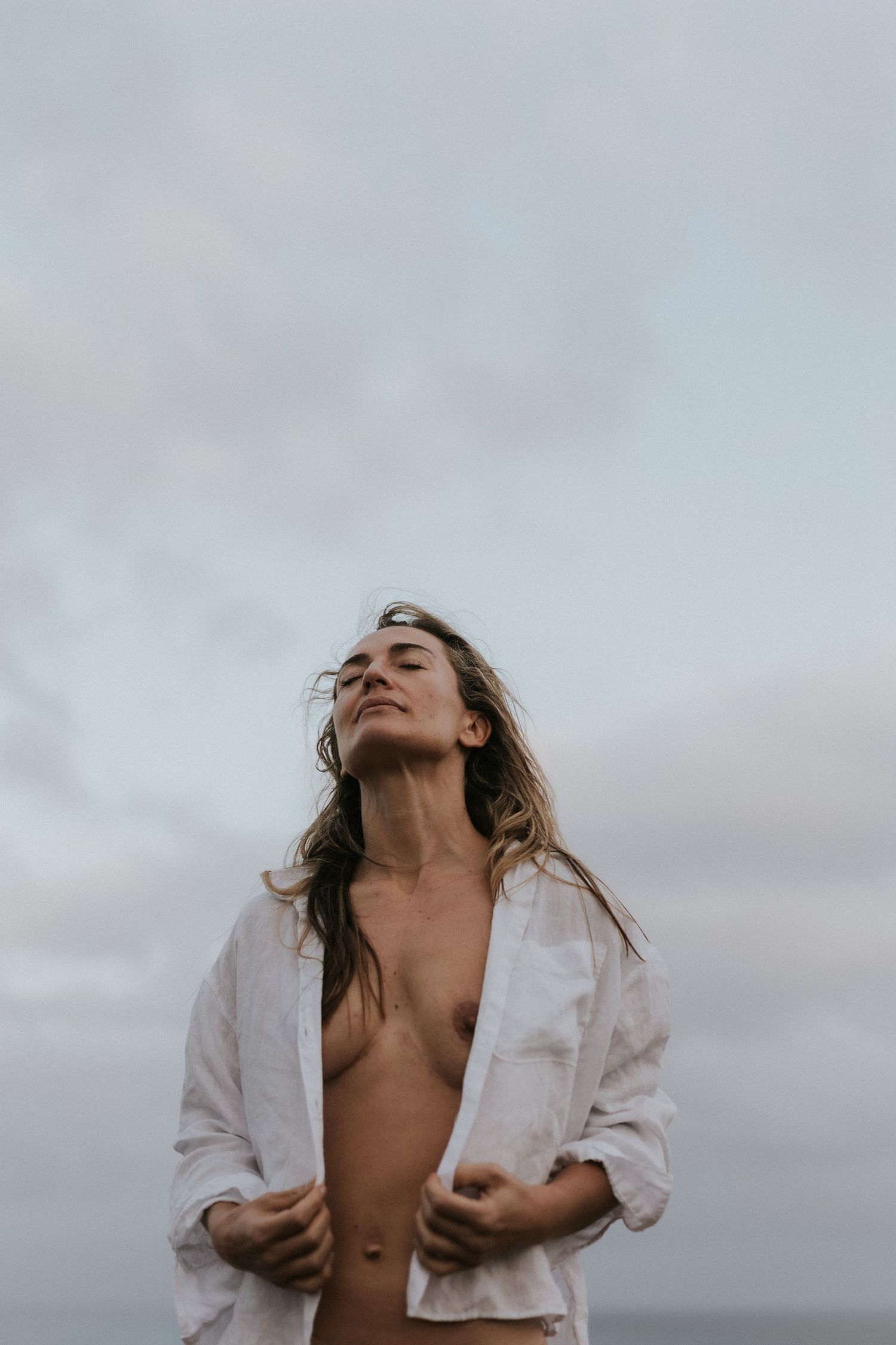 Woman in open white linen shirt standing outdoors against cloudy sky with eyes closed and wind-blown hair.