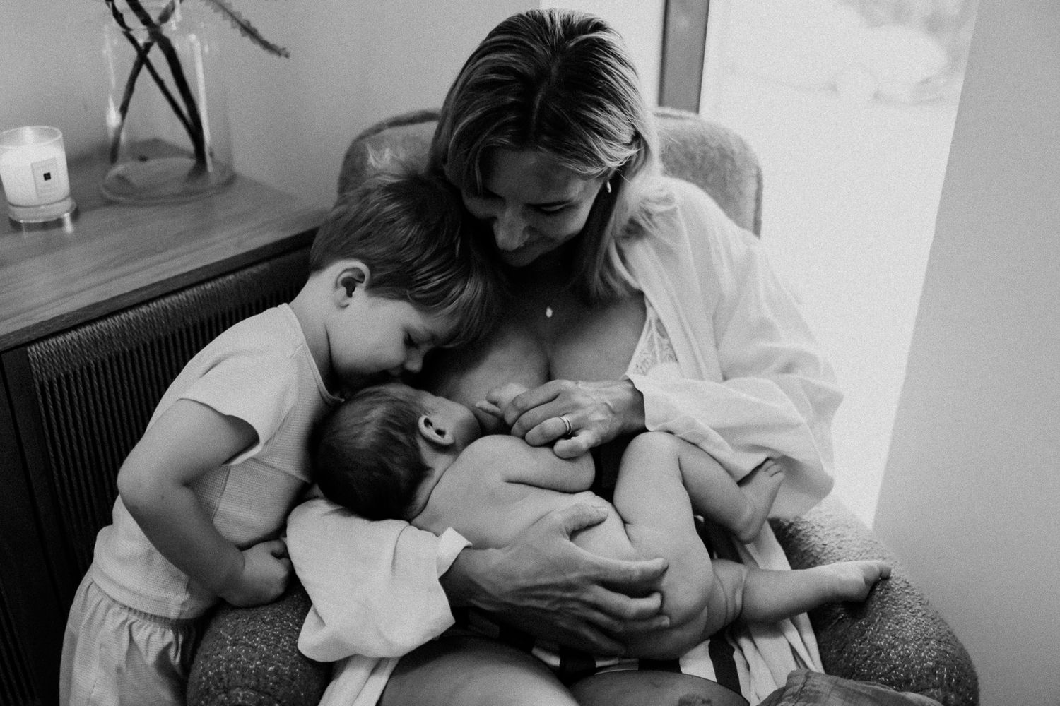 Black and white photo of a mother breastfeeding newborn while older child cuddles close in a serene home setting.