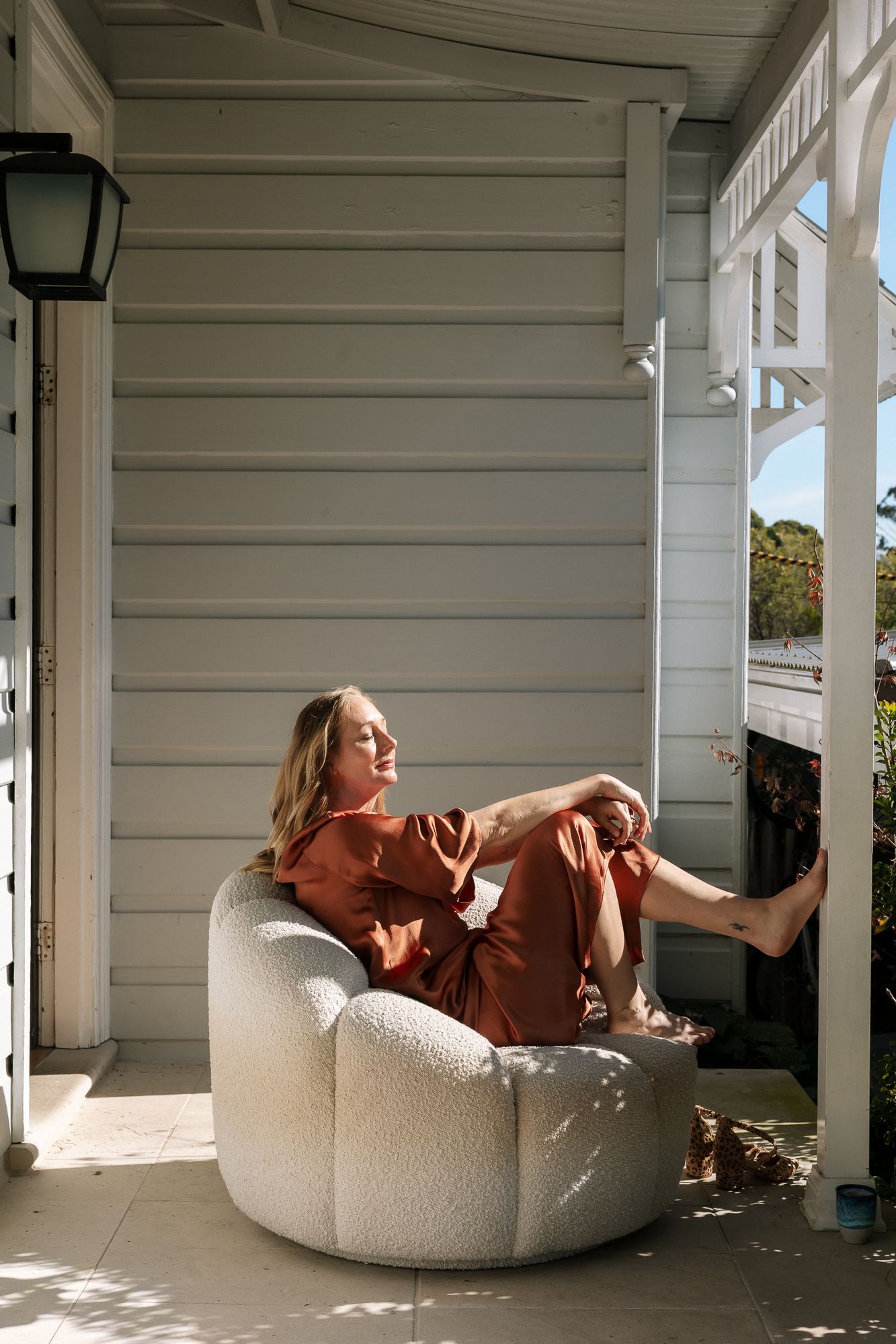 Woman in rust-colored linen set relaxing on a white boucle chair on a sunlit white timber porch.