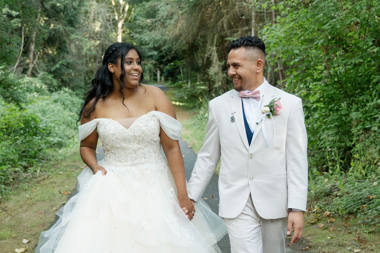 Bride in white off-shoulder gown and groom in light gray suit hold hands smiling on a lush green forest path.