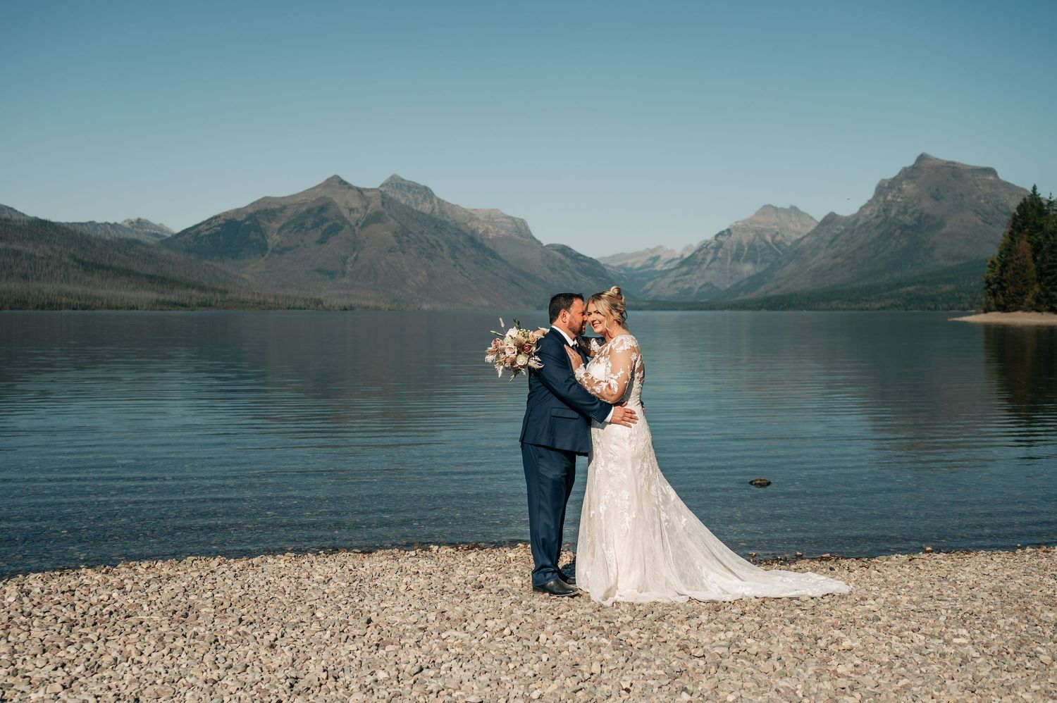 Bride and groom kissing on rocky lakeshore with stunning mountain backdrop under clear blue sky.