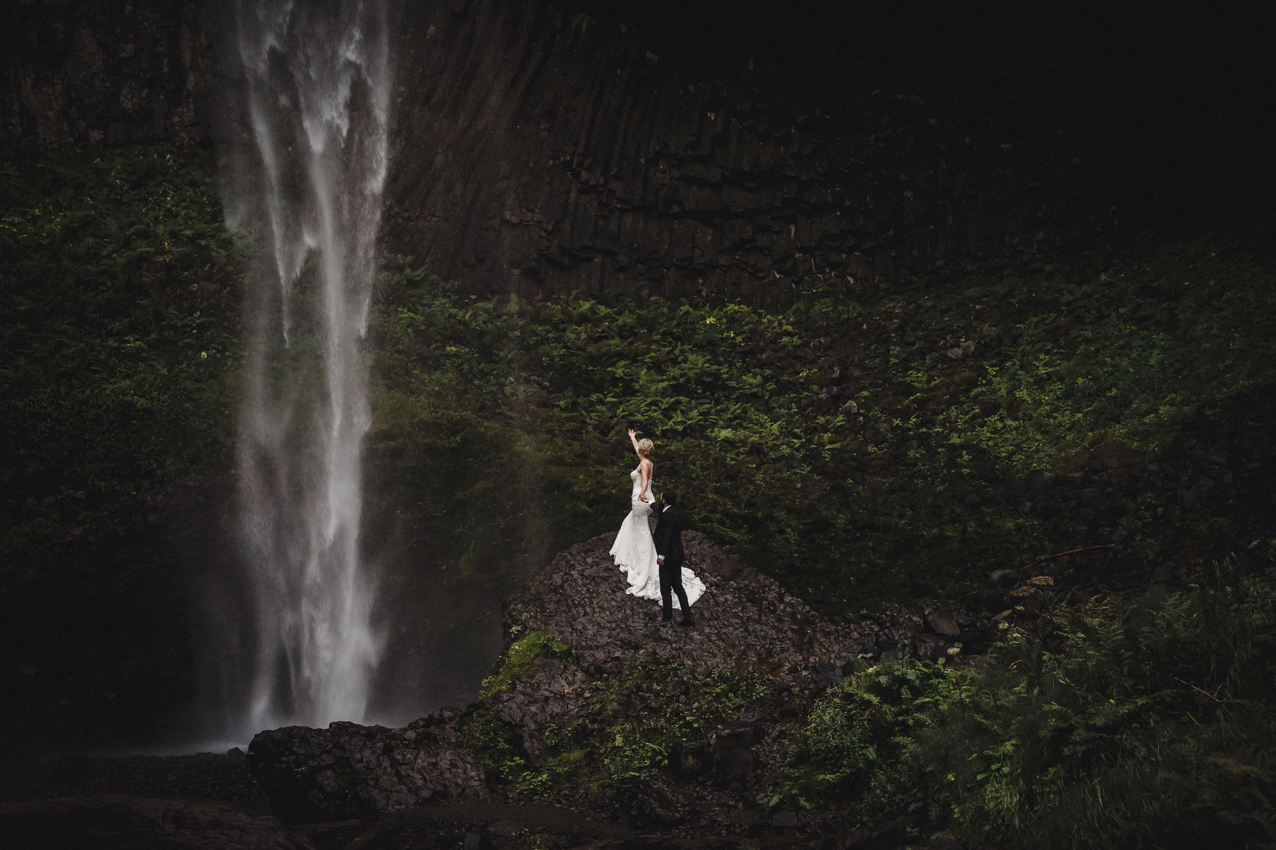 A bride and groom stand romantically on rocks before a majestic waterfall surrounded by lush green foliage.