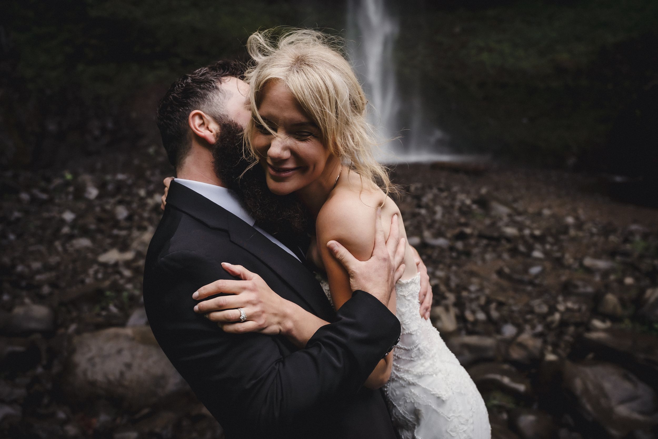 A couple embraces joyfully in wedding attire before a lush waterfall backdrop on their special day.