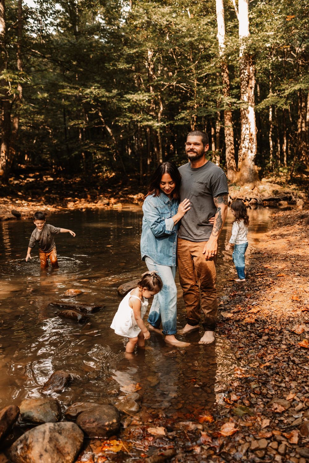 Family of four playing in a forest creek on a sunny autumn day, parents embracing while children explore the water.