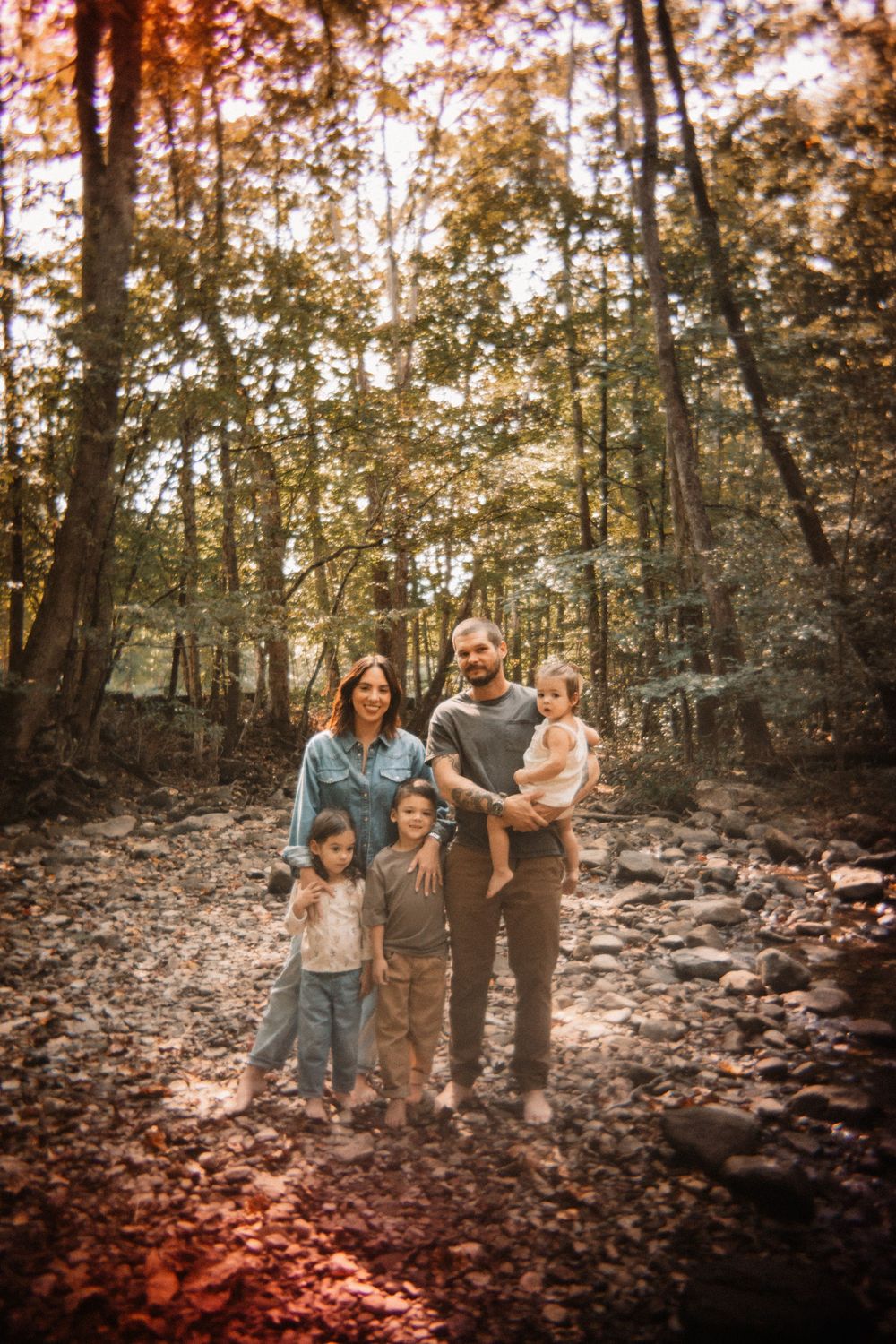 Family of five posing together in a sunlit autumn forest with rocky ground and tall trees.