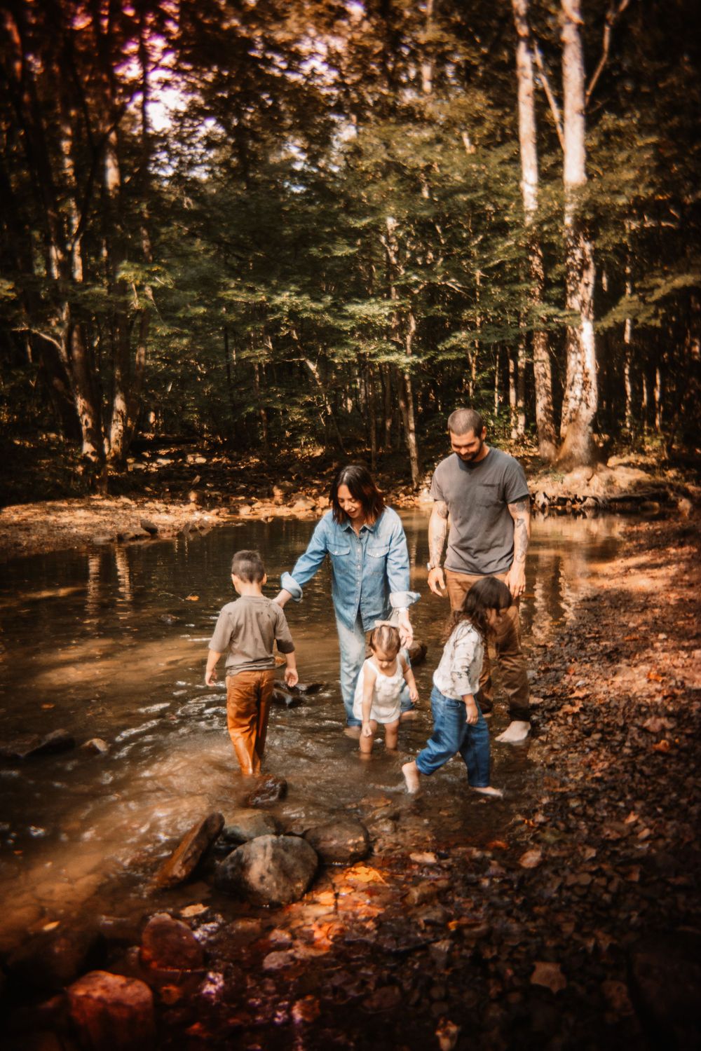 Family of five walking together through shallow creek in autumn forest.