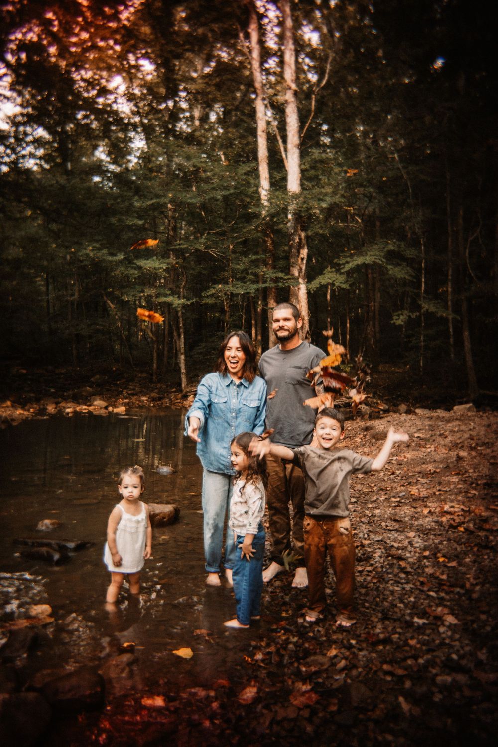 Family of five laughing and tossing autumn leaves by forest creek.