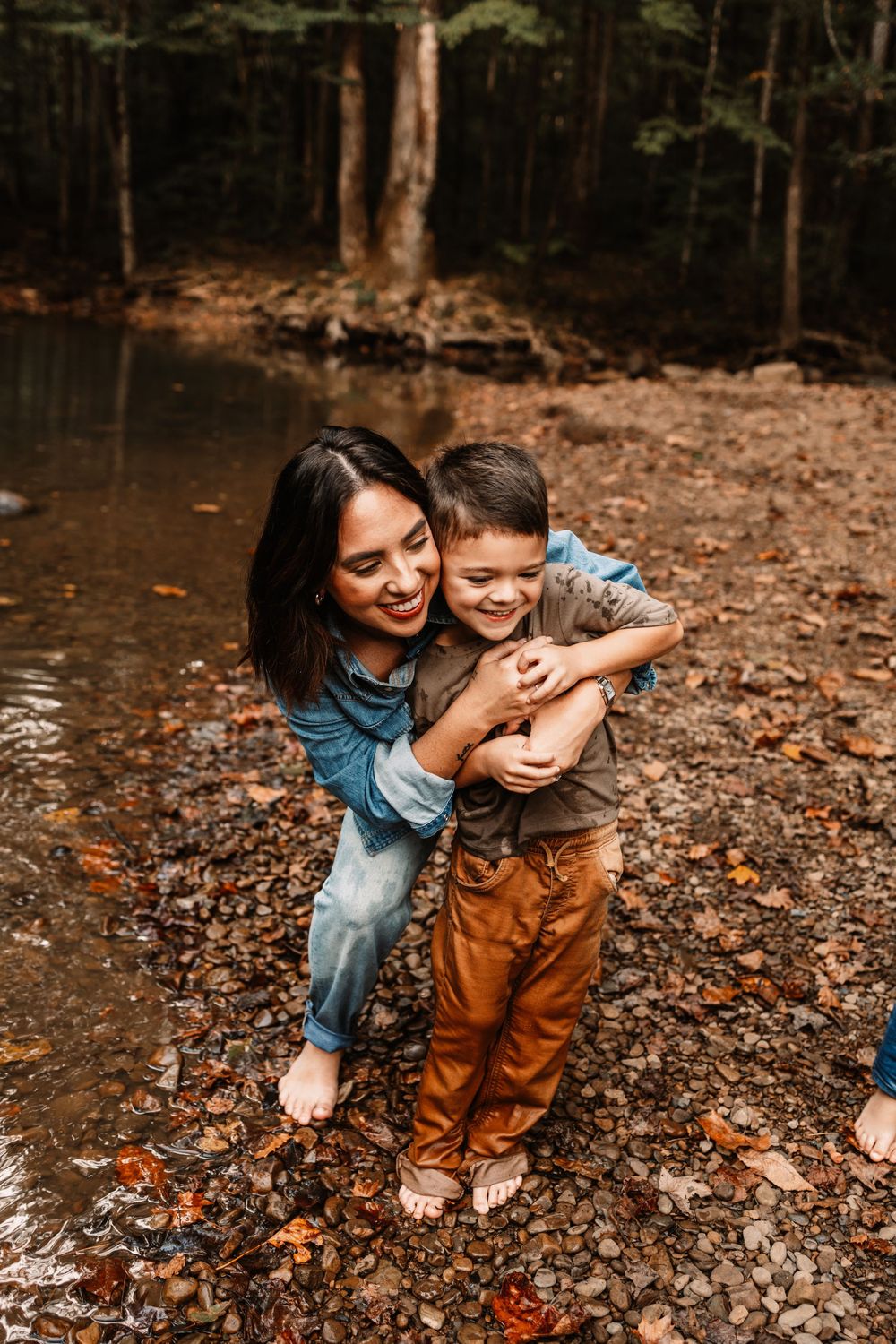 A smiling mother hugs her young son outdoors near a forest creek surrounded by autumn leaves.