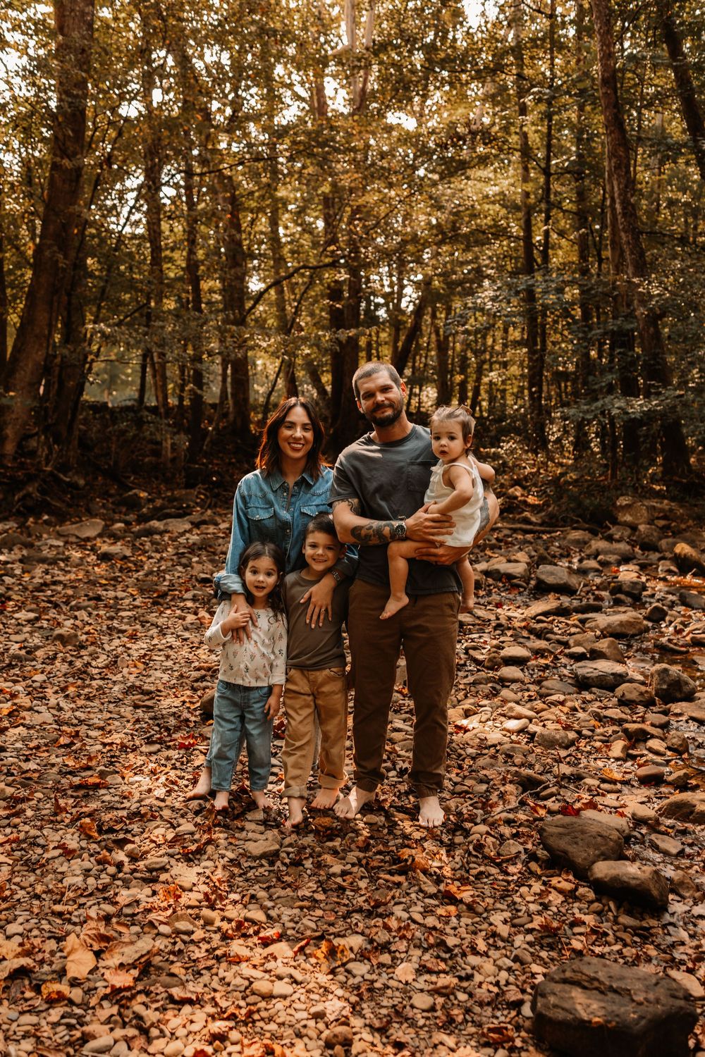 Family of five posing in autumn forest with fallen leaves, two parents and three young children smiling outdoors.