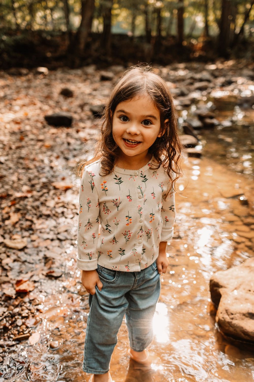 A smiling young girl in a floral top and jeans stands barefoot in a shallow woodland creek.