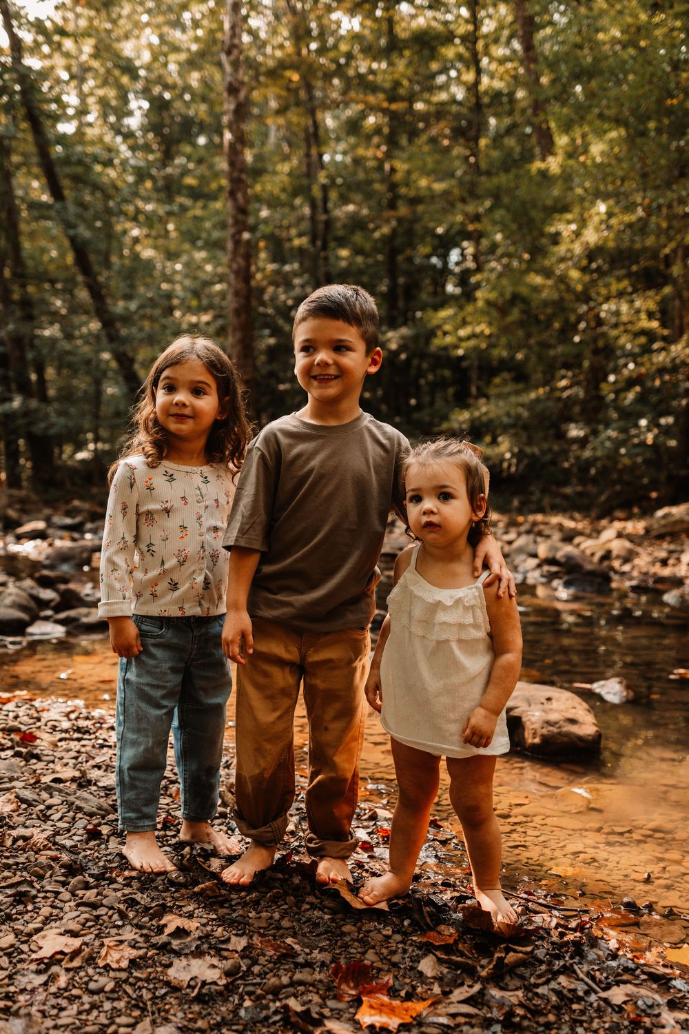 Three barefoot children pose by a forest creek surrounded by autumn leaves and lush green trees.