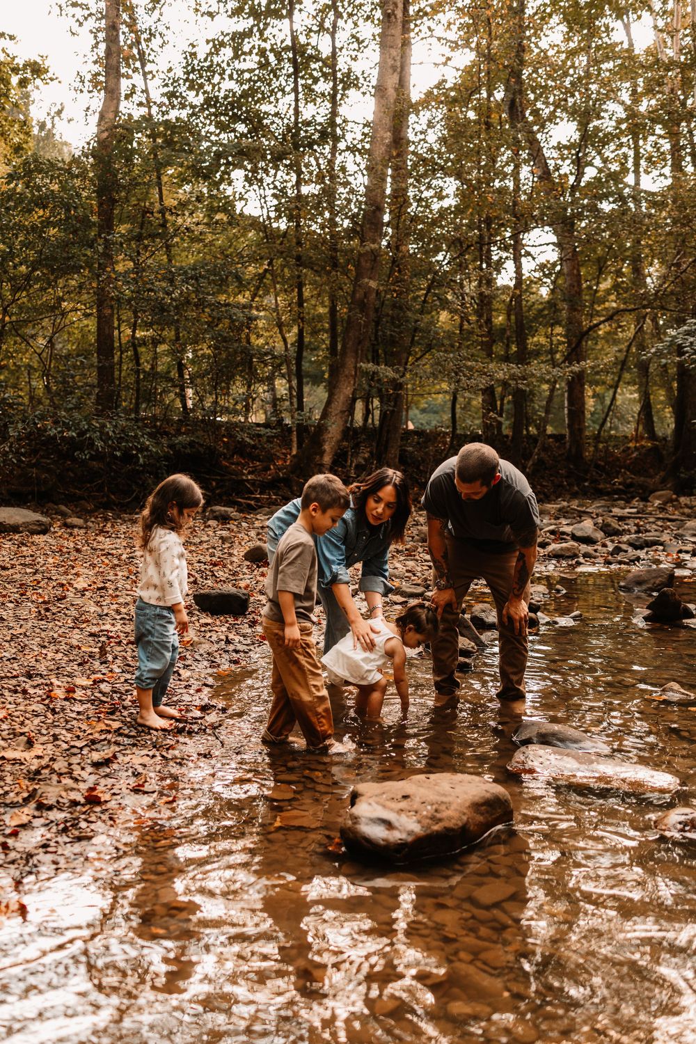 Family of four playing in a shallow forest creek surrounded by autumn leaves and tall trees.