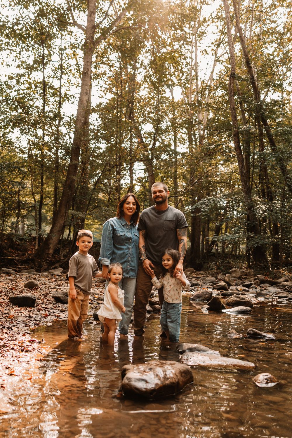 Family of five smiling together in a sunlit forest creek, surrounded by lush green trees and rocky terrain.
