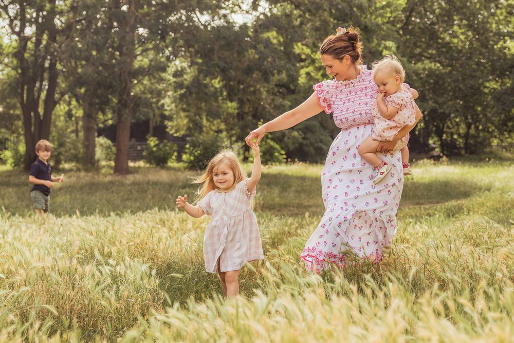 A mother in a floral dress plays with her children in a sunny green meadow.