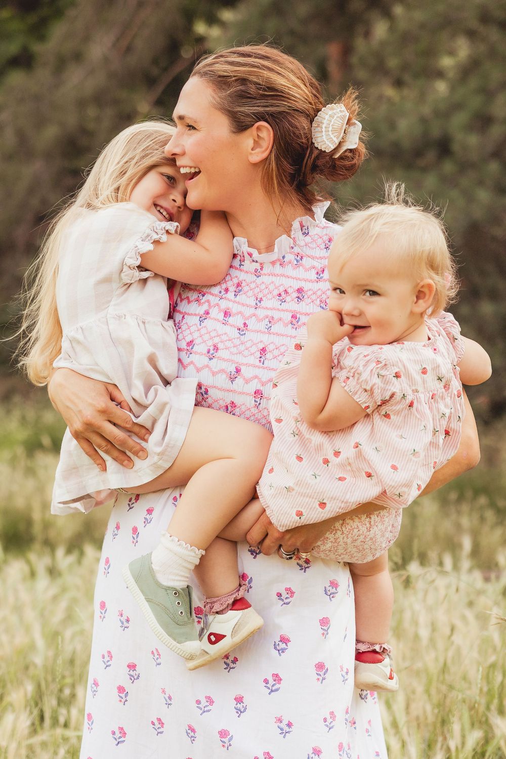 A smiling mother in a floral dress holds two young blonde girls outdoors in a lush green field.