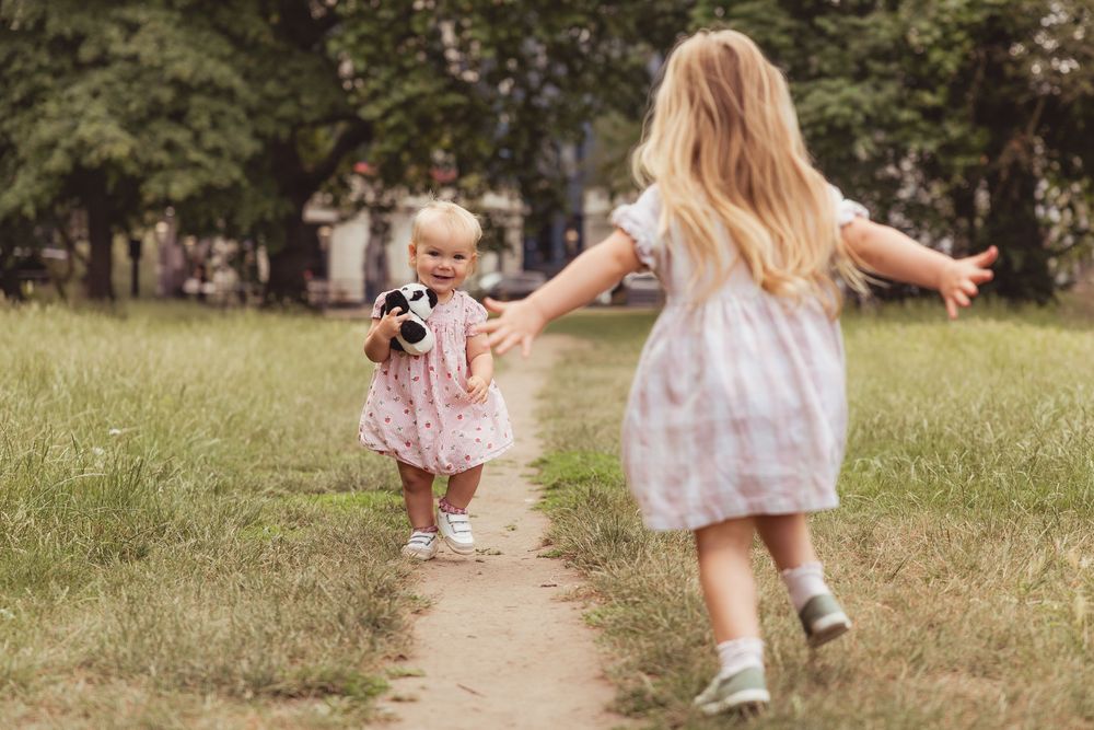 Two young girls in dresses playing on a dirt path in a lush green park, one holding a toy camera.