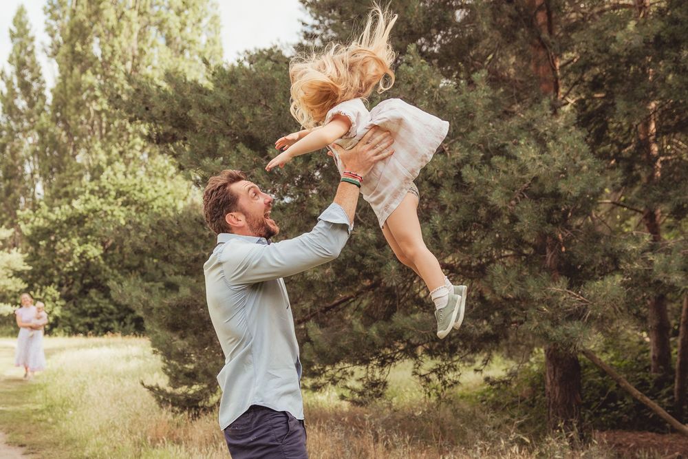 A joyful father tosses his laughing little girl into the air in a sunny green park setting.