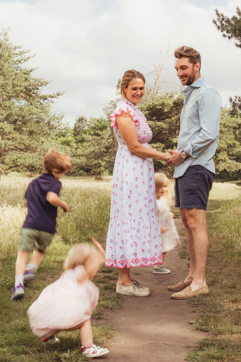 Happy couple holding hands on a park path while young children play around them on a sunny day.