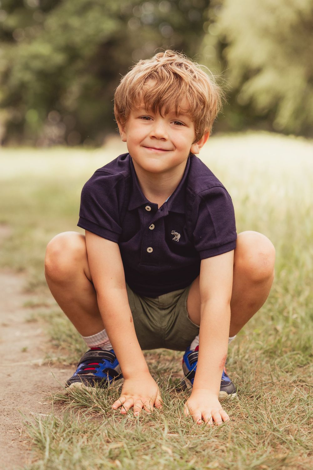 A young smiling boy in a navy polo shirt crouches on grass in a sunny outdoor park setting.