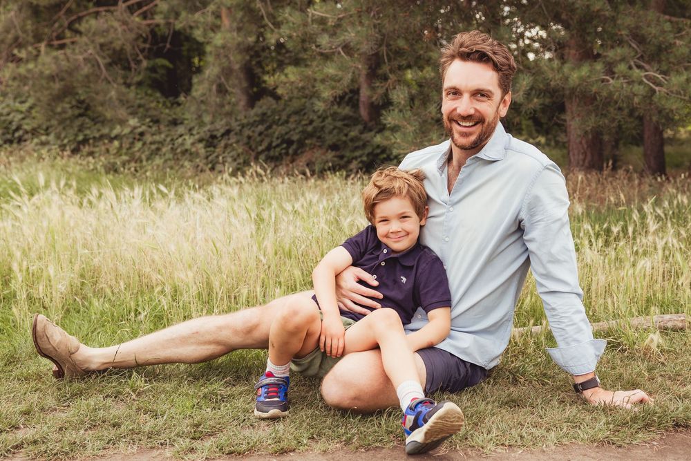 A smiling father and young son sit together on grass near tall reeds and trees in an outdoor family portrait.