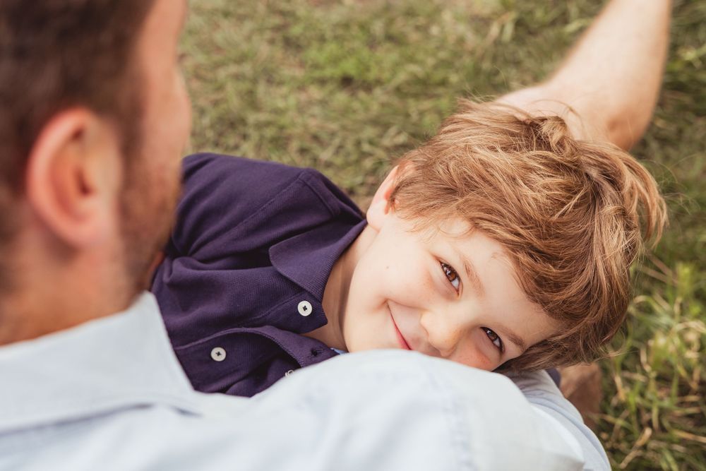 A young smiling boy rests his head on a man's chest while lying on green grass outdoors.