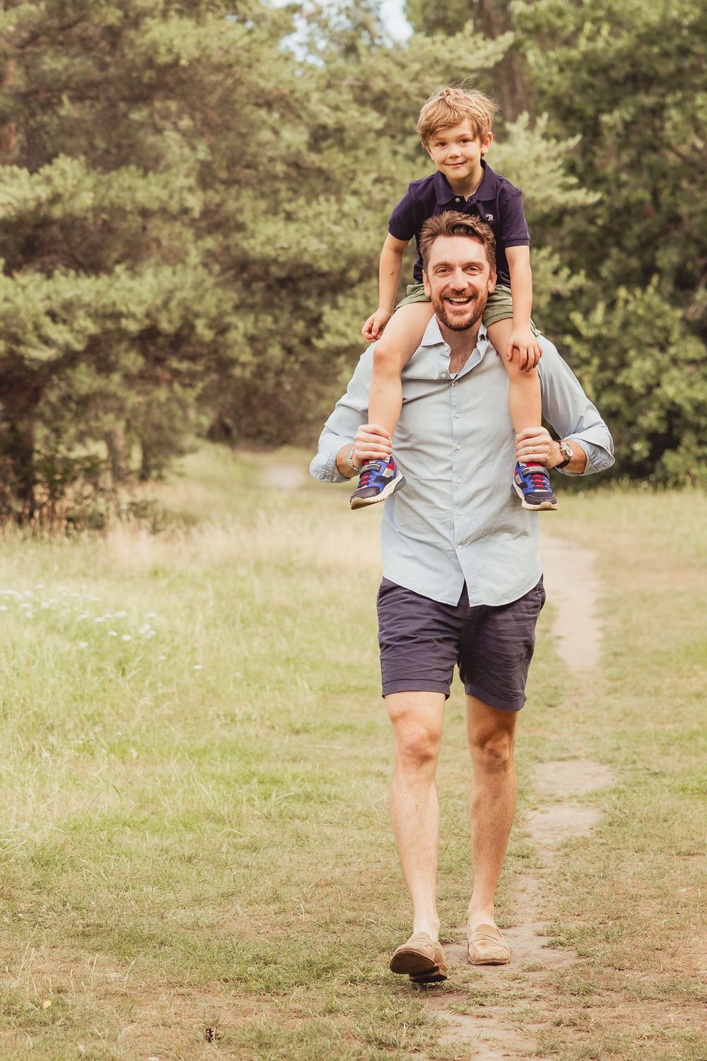 A laughing father carries his young son on his shoulders along a sunny park trail.