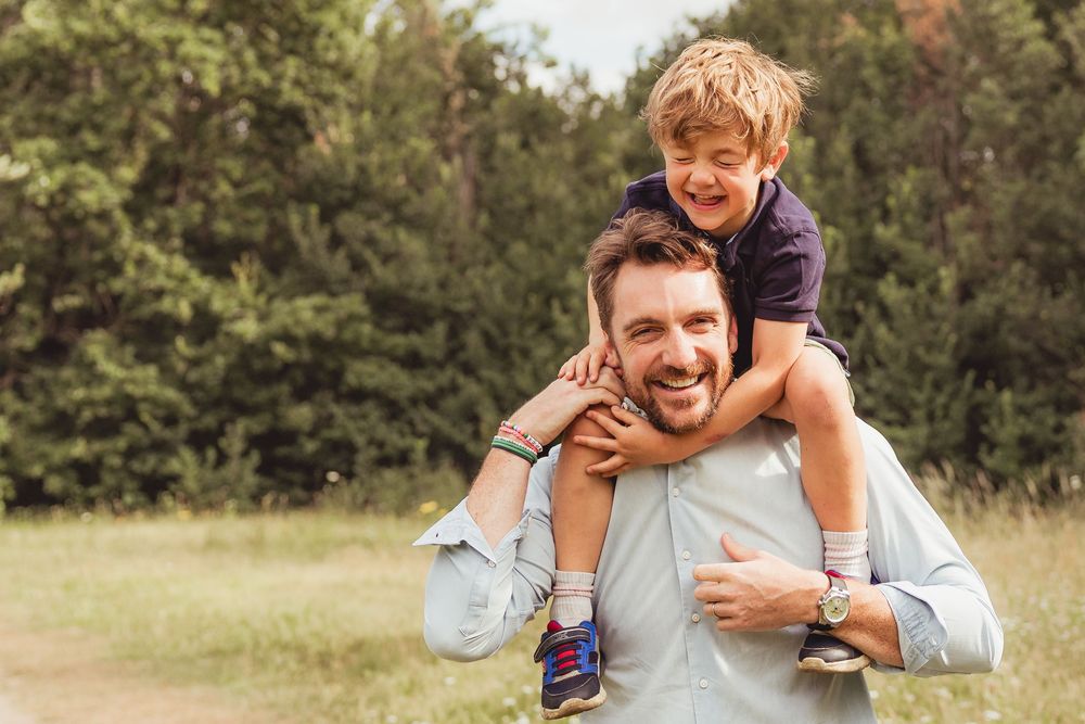 Happy father carrying smiling young son on his shoulders outdoors in a green park setting.