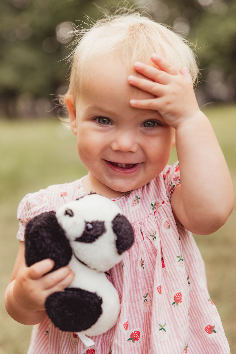 Smiling toddler girl in strawberry dress holding panda stuffed animal outdoors.