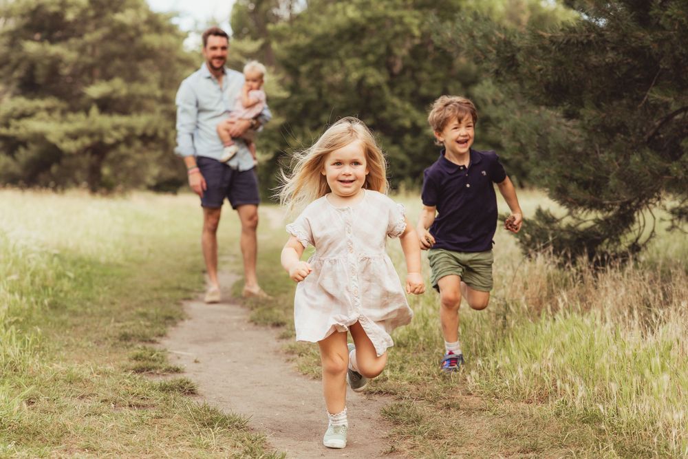 A father holds a baby while two children run along a path in a lush park.