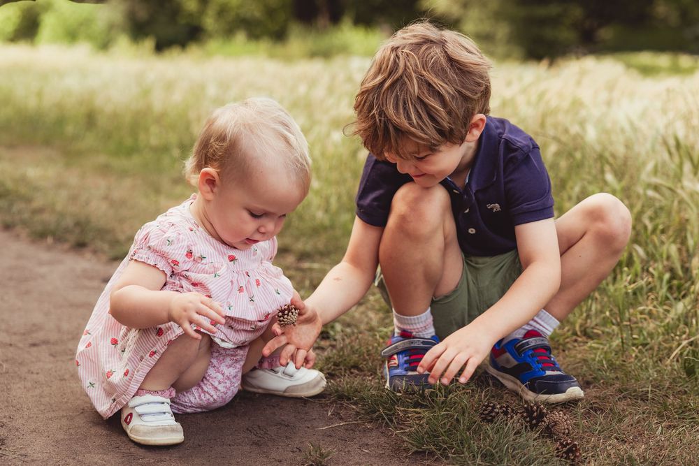 A young boy and toddler girl explore the ground together on a sunny dirt path.