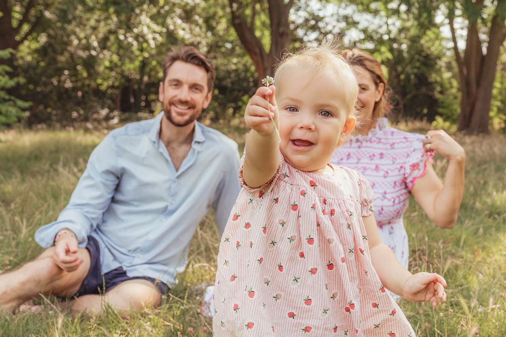 Happy family with smiling parents sitting on grass while cute toddler girl in pink dress holds a flower outdoors.