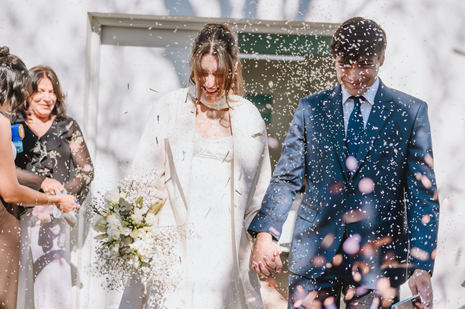 Bride and groom exit church ceremony as guests toss white confetti, bride holding bouquet in elegant white coat.