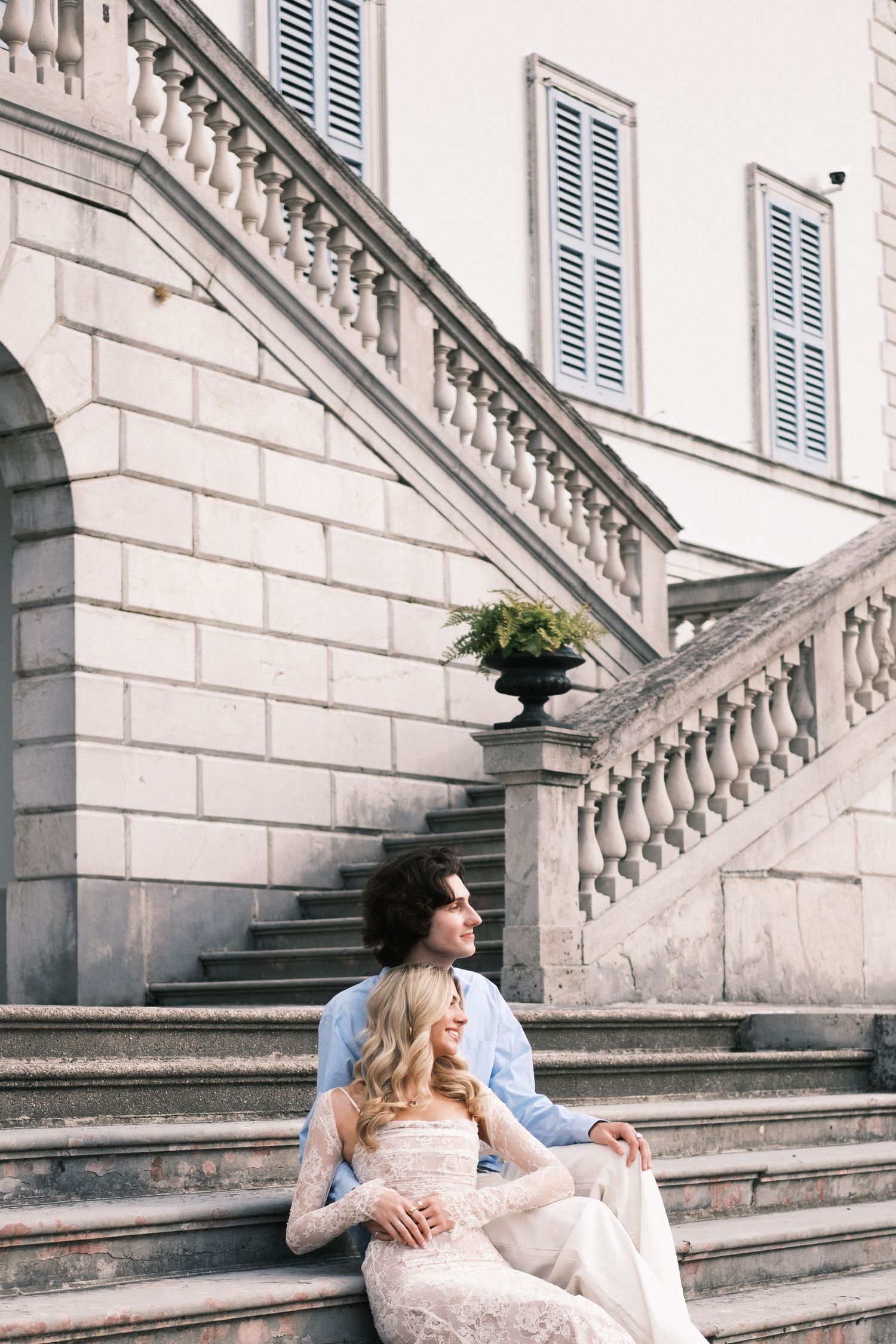 A bride and groom sit together on elegant stone steps of a classic European building, sharing a romantic moment.