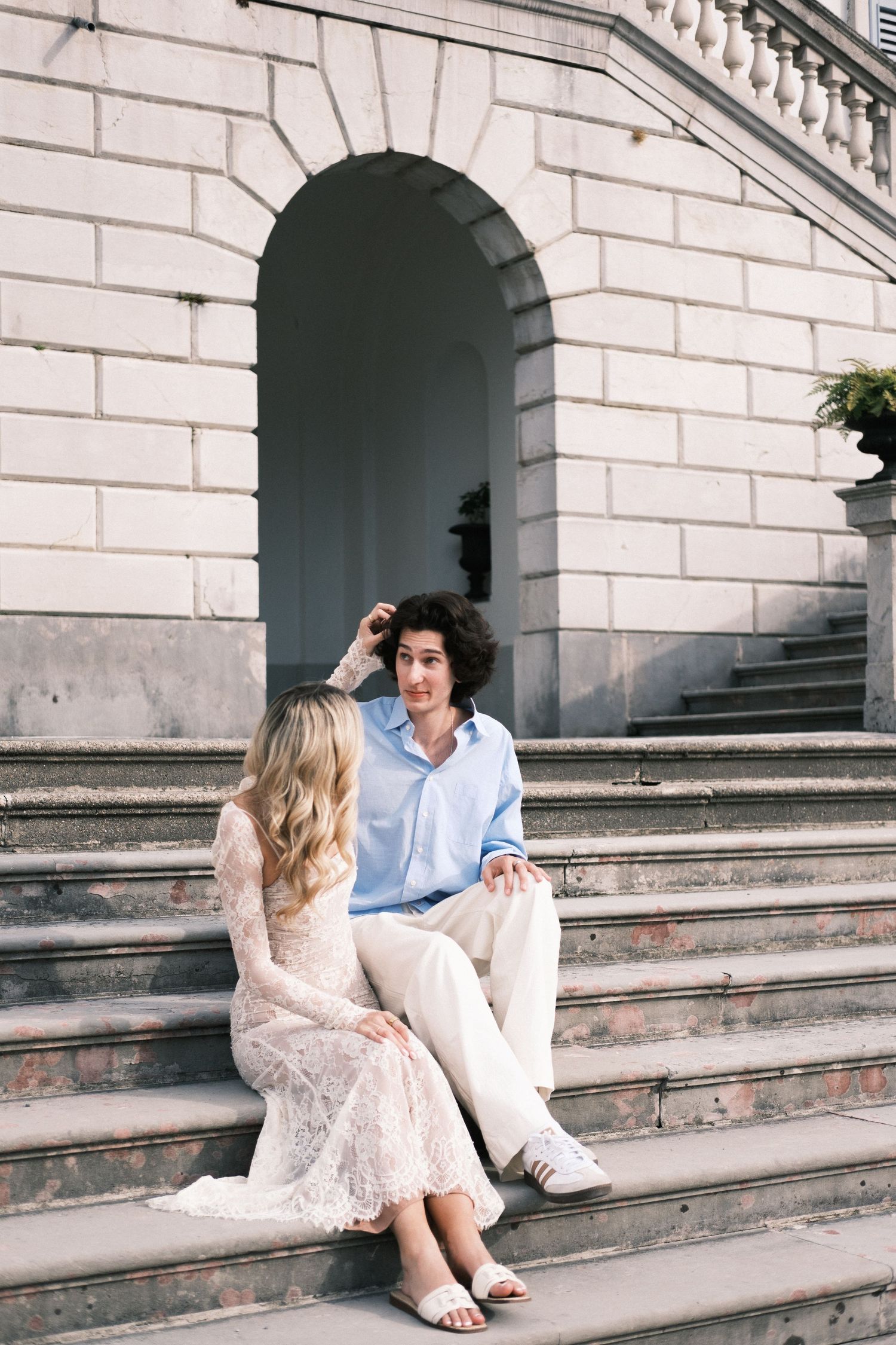 A young couple sits on stone steps of a classical building, wearing elegant casual attire, engaged in intimate conversation.