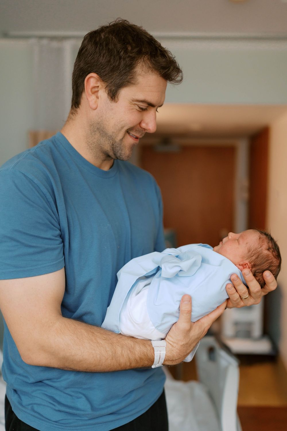 Proud new father in blue shirt holding swaddled newborn baby in hospital room.