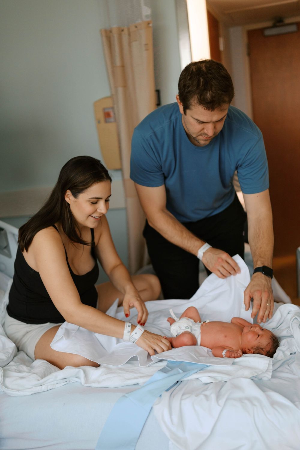 Smiling new parents in hospital room carefully unwrapping their swaddled newborn baby on bed.