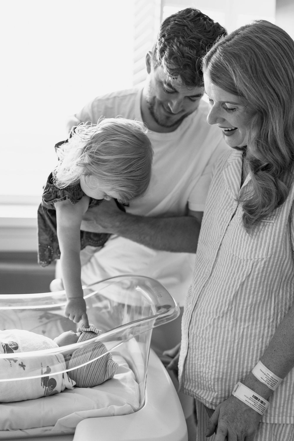 Black and white photo of parents and toddler admiring newborn baby in hospital bassinet.