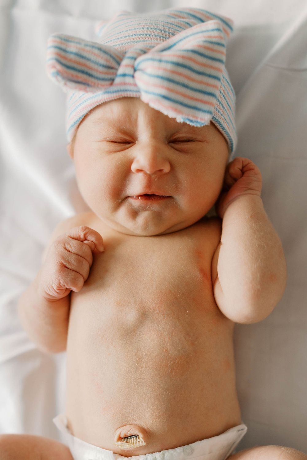 Newborn baby in striped bow hat lying on white blanket, scrunching face with tiny fists raised.