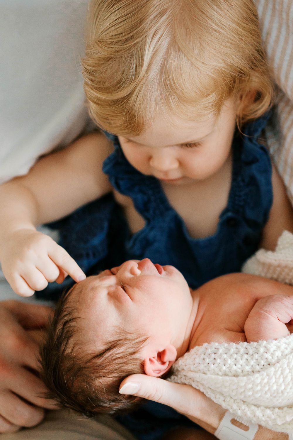 A blonde toddler in blue dress gently touching a sleeping newborn baby held in adult arms.