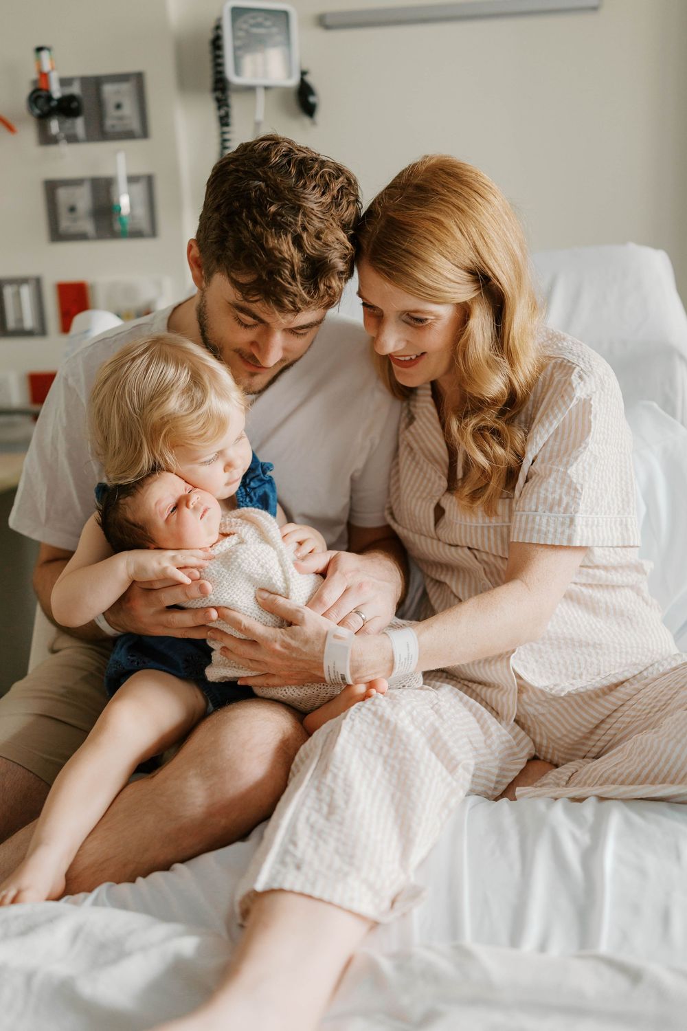 New parents and toddler sitting on hospital bed, lovingly gazing at swaddled newborn baby.