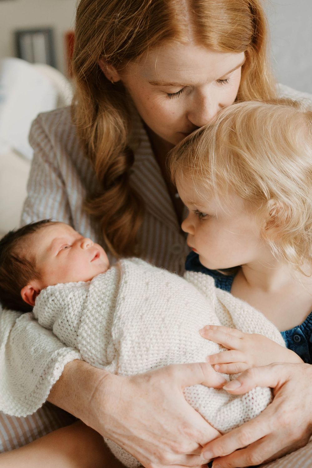 A red-haired mother kisses her toddler while holding a sleeping newborn baby wrapped in white knit.