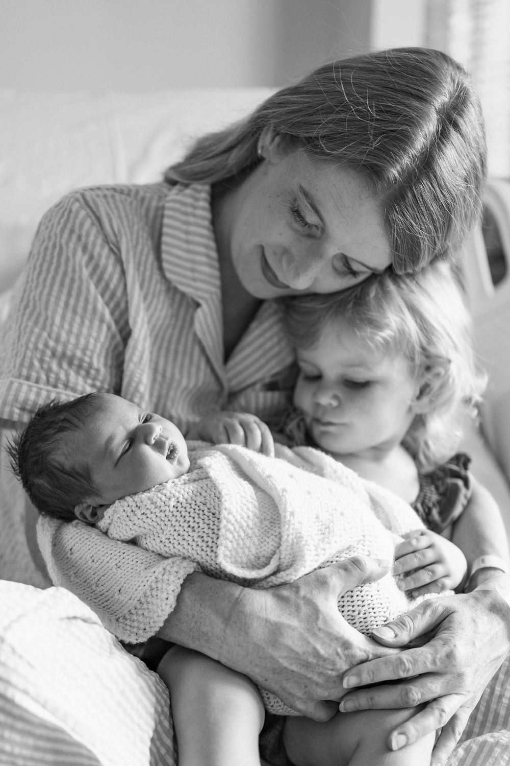 Black and white photo of a mother holding newborn baby with toddler sibling on hospital bed.