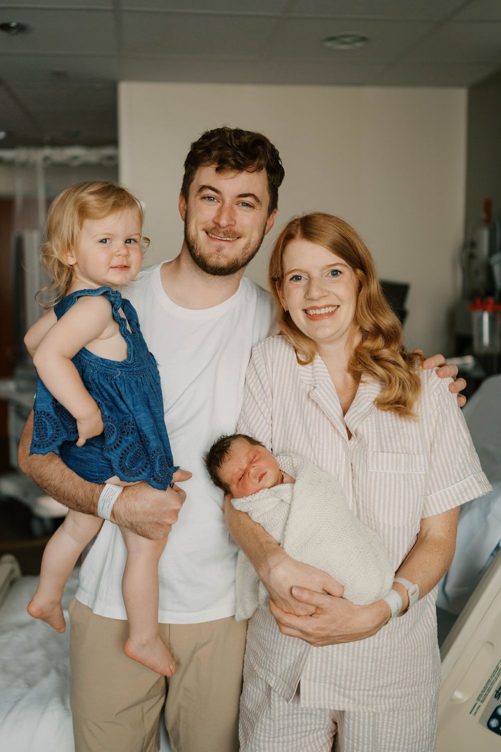 A smiling family of four poses in a hospital room with toddler and swaddled newborn baby.