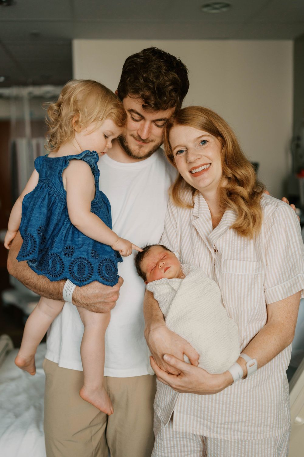 Happy family of four in hospital room, parents holding newborn baby and toddler girl in blue dress.