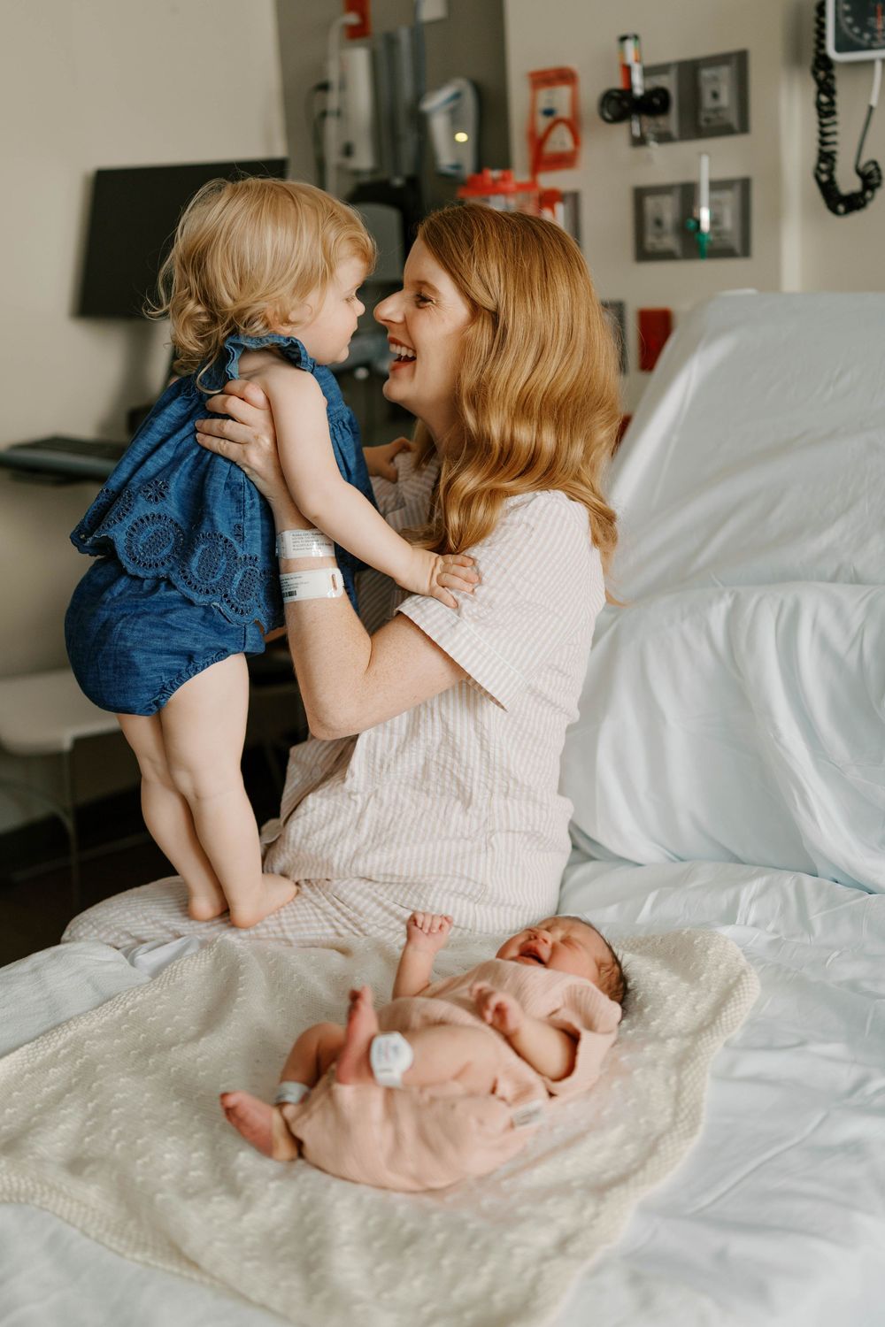 A mother on a hospital bed lifts her toddler while newborn baby rests on the bed beside her.