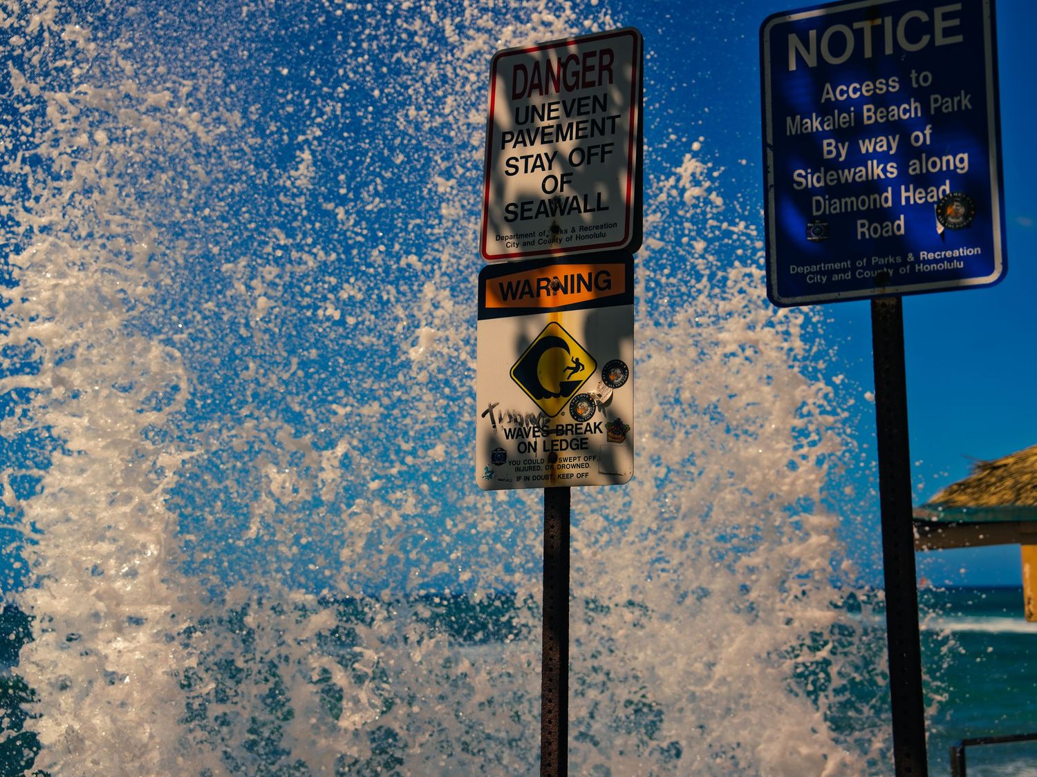 A wave crashes into a huge spray of white water behind a seawall danger sign and a Makalei Beach Park notice sign in Honolulu, Hawaii.