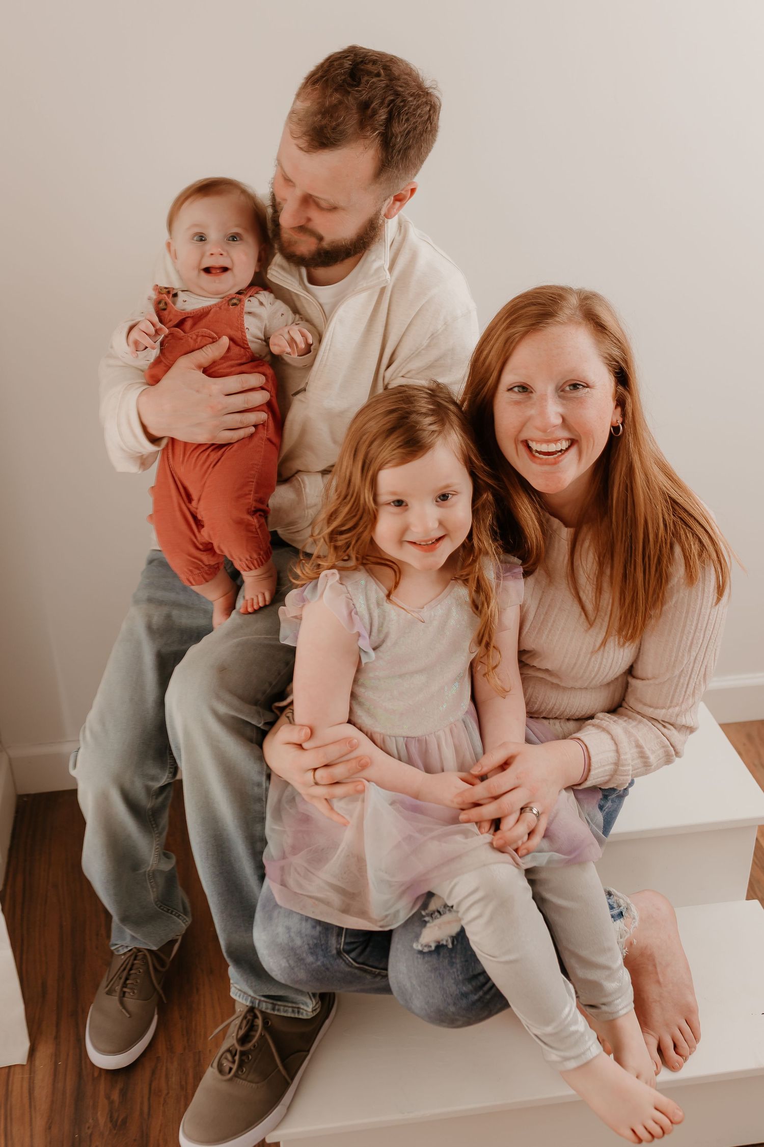 Happy family of four posing for a portrait, parents with two young daughters, smiling in a bright white studio setting.