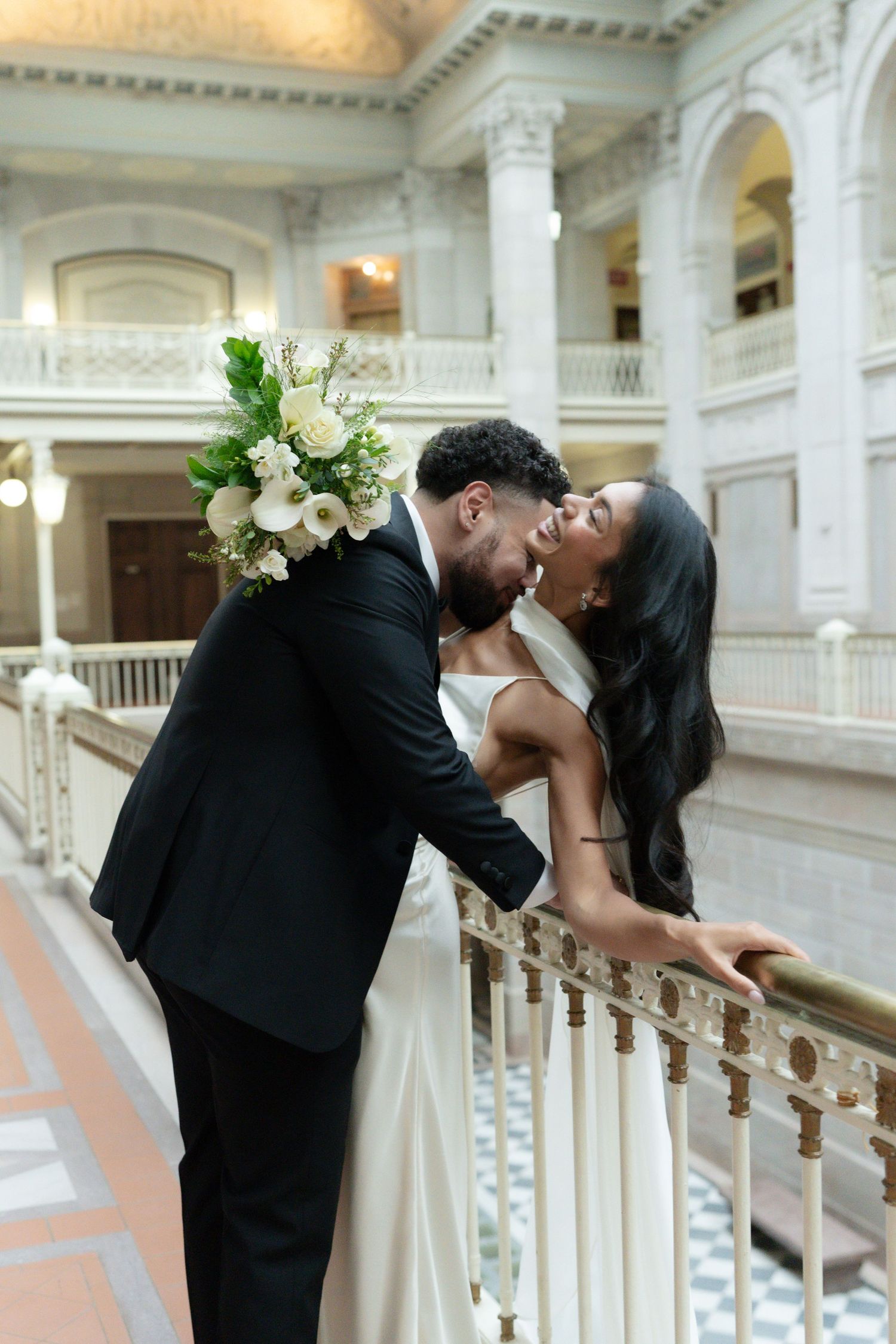 Bride and groom sharing a romantic kiss on a balcony railing inside an elegant neoclassical venue.