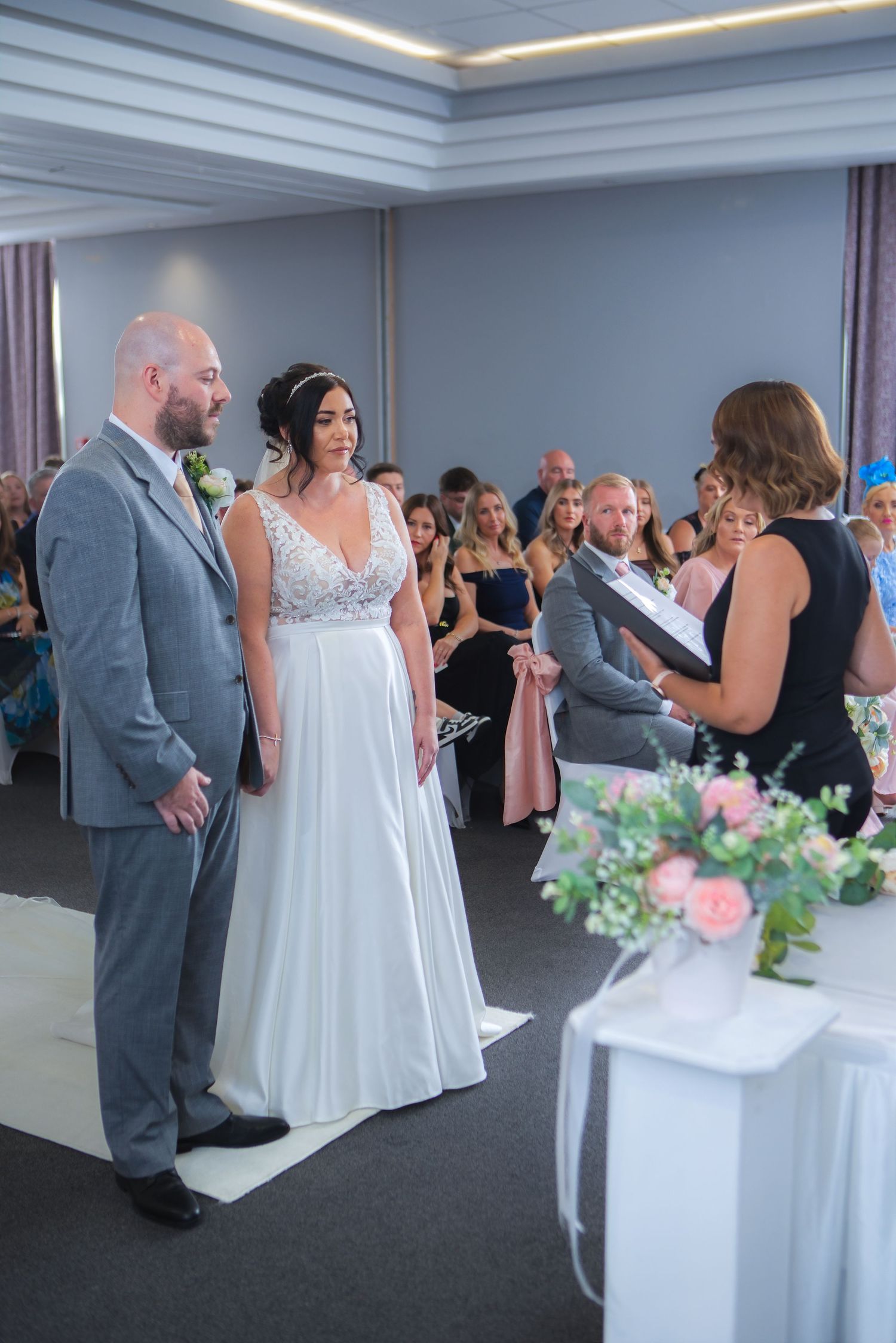 Bride and groom exchange vows at indoor wedding ceremony with guests seated in background.