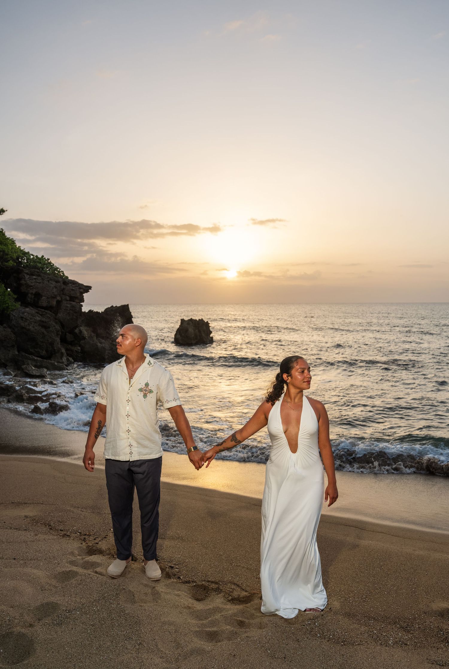Bride and groom hold hands on a sandy beach at sunset with ocean waves and rocky cliffs in the background.